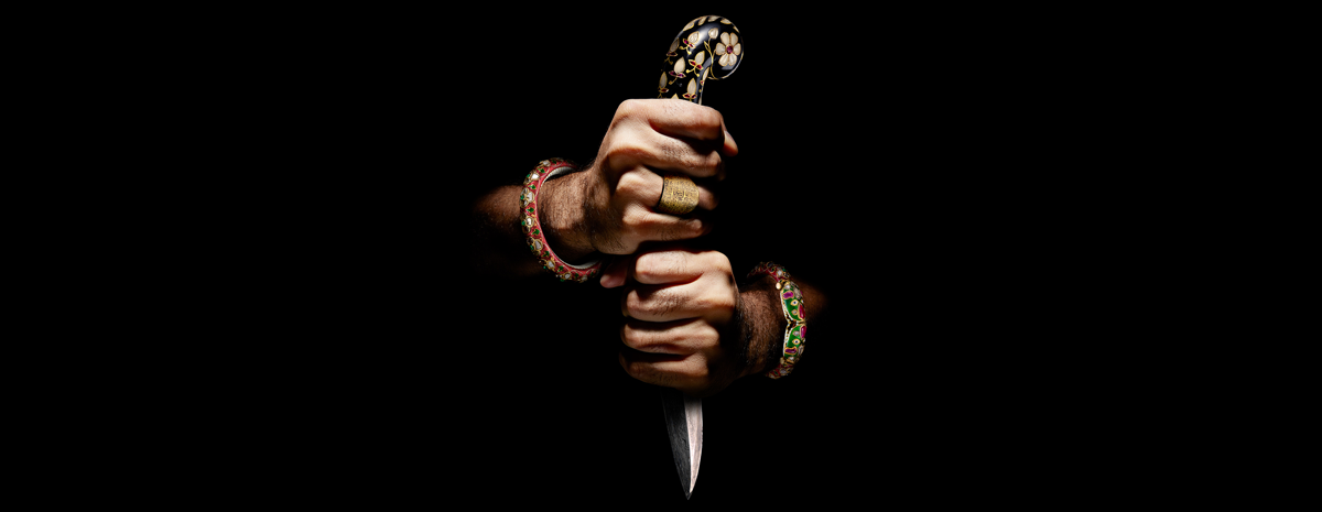 Two hands decorated in colourful jewellery emerging from darkness. The hands are tightly gripped around an opulent dagger.